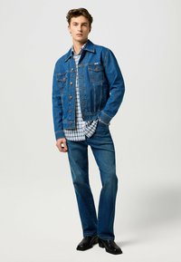 Veste en denim avec fermeture à boutons, poches poitrine et coutures contrastantes, associée à une chemise à carreaux et un jean bleu, le tout complété par des bottes noires.