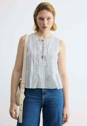 Mladá žena s krátkymi blond vlasmi oblečená v bielej bezrukávovej blúzke s viazaním vpredu, v modrých džínsoch a s béžovou kabelkou cez rameno.