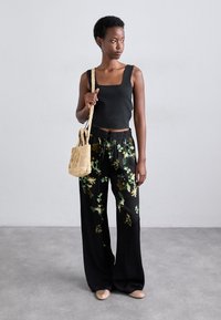 Černé přiléhavé tílko spojené s širokými černými kalhotami s květinovým vzorem v zelených a žlutých odstínech. Drží malou pletenou slaměnou tašku.
