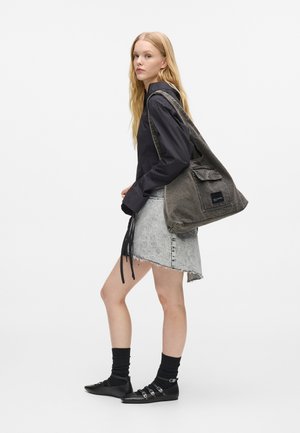 Νεαρή γυναίκα στέκεται, φοράει μαύρη μπλούζα με μακριά μανίκια, ανοιχτόχρωμη τζην φούστα, μαύρες κάλτσες με παπούτσια με πόρπες, και κρατάει μια μεγάλη γκρι τσάντα ώμου.