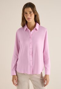 Femme aux longs cheveux bruns portant une chemise rose pâle à boutons et un pantalon beige, debout devant un fond beige uni.