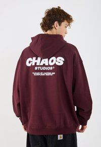 Hanorac vișiniu cu un imprimeu alb mare care spune "CHAOS STUDIOS" pe spate. Are un glugă cu șnur și manșete ribbate.