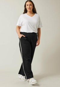 Pantaloni larghi neri con strisce laterali bianche, abbinati a una semplice t-shirt bianca a V e sneaker bianche. Il tessuto appare morbido.