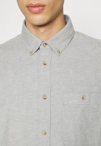 Camisa gris de botones con cuello, que presenta botones marrón claro y un bolsillo en el pecho. La tela es suave con una textura sutil.