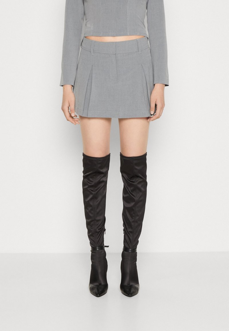 Femme portant une mini-jupe plissée grise et des bottes noires à talons au-dessus du genou, debout devant un fond blanc.