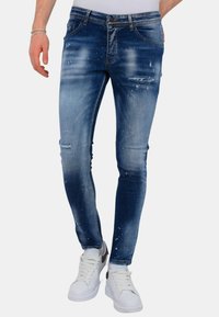Man in slim-fit blauwe versleten jeans en witte sneakers met zwarte hakdetails, staande tegen een effen achtergrond.