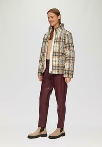 Geruite gewatteerde jas in crèmekleurige en bruine tinten, gecombineerd met bordeauxkleurige neplederen broek en beige enkellaarsjes met zwarte accenten.