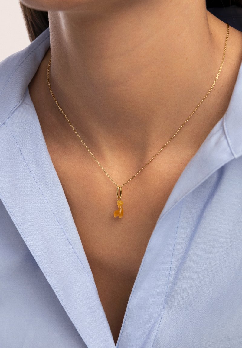 Eine goldene Halskette mit einer zarten Kette und einem gelben Edelstein-Anhänger in Form eines Fisches, getragen über einem hellblauen Hemd.