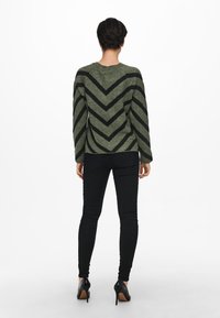 Pull vert avec des rayures chevrons noires, tricot texturé, col rond et manches longues, associé à un jean noir slim et des talons noirs.