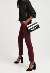 Blouse noire, pantalon bordeaux et chaussures à talons noires. Le sac à main présente des rayures noires et blanches avec un détail de fermoir doré.