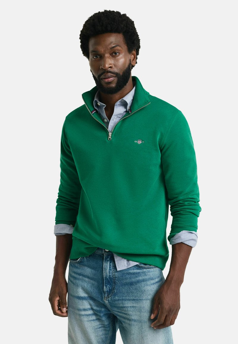 Grön zip-up sweatshirt med hög krage, stickad textur och logotyp. Bärs över en ljusblå skjorta med knappar, i kombination med urtvättade blå jeans.