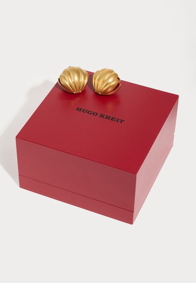 Χρυσά σκουλαρίκια σε σχήμα κοχύλι που ακουμπούν σε ένα κόκκινο κουτί με την επιγραφή «HUGO KREIT» ανάγλυφη σε μαύρο. Το κουτί έχει λείο, ματ φινίρισμα.