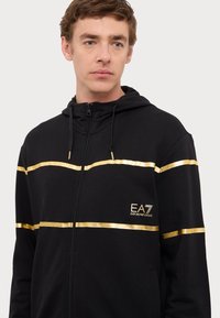 Black zip-up hoodie featuring gold stripes across the chest and left side. Material is soft with a textured finish; includes drawstring hood.