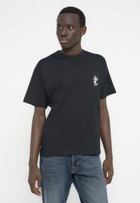 Μαύρο βαμβακερό t-shirt με κοντά μανίκια, λαιμόκοψη με ρέλι και μικρό γραφικό σχέδιο στο αριστερό στήθος. Συνδυασμένο με μπλε τζιν παντελόνι.