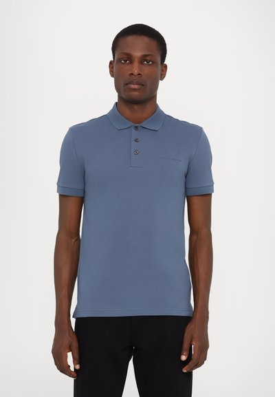 Tricou polo de culoare albastră deschisă, realizat dintr-un material neted. Dispune de un guler, două rânduri de butoni pe față și mâneci scurte. Design simplu.