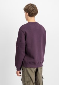 Ljubičasti dukserica s okruglim izrezom, rebrastim manžetama i donjim rubom. Sadrži teksturiranu zakrpu na lijevom rukavu i mali detalj s logotipom.