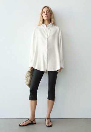 Kobieta ubrana w oversize'ową białą koszulę zapinaną na guziki, czarne krótkie legginsy, czarne sandały na paski, trzymająca plecioną kopertówkę na tle gładkiej ściany.