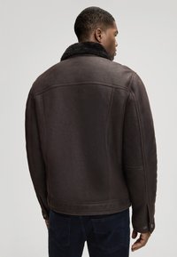 Brązowa skórzana kurtka z miękkim czarnym kołnierzem z futra, charakteryzująca się strukturalnym krojem, widocznym przeszyciem i lekko teksturowanym wykończeniem.