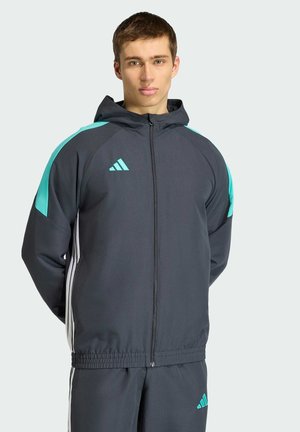 Młody mężczyzna w ciemnoszarym i morskim dresie Adidas z kapturem, stoi zwrócony przodem na jasnym tle.