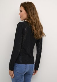 Crni longsleeve s dugim rukavima s valovitim izrezanim uzorcima; teksturirana tkanina; uski kroj; uparen s plavim trapericama; minimalistički, ležeran izgled.