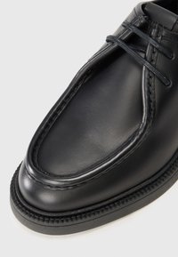 Zapato de cuero negro con punta redondeada, detalles de costura y diseño con cordones. La suela es texturizada con un borde elevado.