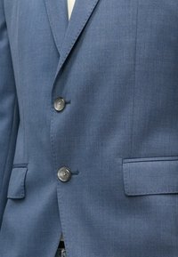 Μπλε σακάκι από υφασμάτινη υφή με δύο μεταλλικά κουμπιά, πέτο με αυλάκι και τσέπη στο στήθος. Ραμμένο σε στενή γραμμή με κομψό σχέδιο.