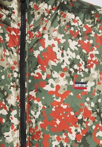 Veste camouflage avec un mélange de motifs verts, rouges et beiges. Comprend une fermeture éclair noire et un petit patch logo Levi's sur la poitrine.