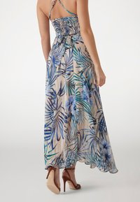 Vestido maxi floral con cuerpo fruncido, que presenta hojas y flores tropicales en tonos azules y verdes sobre un fondo beige. Detalle en la espalda con tirantes.
