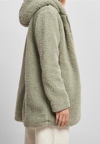 Chaqueta de forro polar verde con una superficie texturizada y esponjosa, capucha y un corte relajado, que llega hasta la mitad del muslo. Diseño sencillo y mangas rectas.