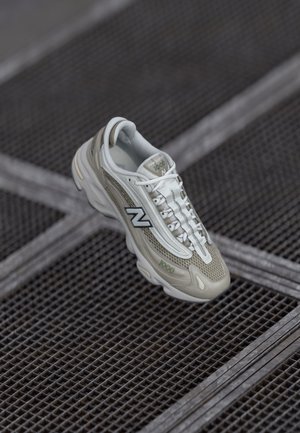 Shoe New Balance gris claro apresenta um cabedal em malha, detalhes texturizados nas laterais e uma sola branca com detalhes em verde e preto.