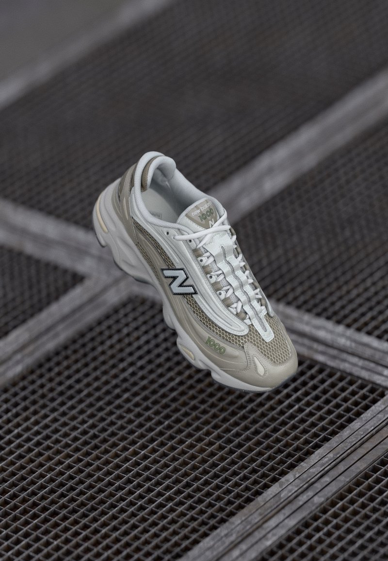 Svetlo šedé tenisky New Balance majú sieťovinový zvršok, textúrované bočné akcenty a bielu podrážku s zelenými a čiernymi detailmi.