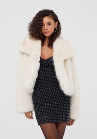 Cappotto in pelliccia ecologica color crema con colletto ampio e maniche piene, abbinato a un vestito nero aderente con pois bianchi e spalline sottili.