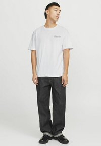 Balta medvilnė t-shirt su trumpomis rankovėmis, turinti mažą spaudinį ant krūtinės, derinama su tamsiai mėlynomis džinsinėmis kelnėmis ir juodais batais.