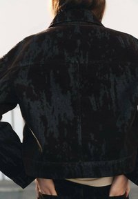 Giacca di jeans nera con superficie testurizzata, caratterizzata da un design cropped, colletto a camicia e sottili variazioni di pattern sul tessuto.