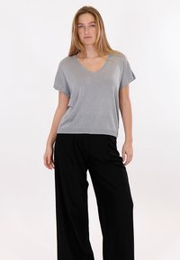 Donna con capelli biondi lunghi, che indossa una maglietta grigia a maniche corte con scollo a V e pantaloni neri larghi, in piedi davanti a uno sfondo bianco.