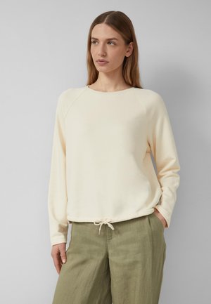 Vrouw draagt een crèmekleurige top met lange mouwen en een trekkoordzoom, gecombineerd met olijfgroene broek met brede pijpen, staand tegen een effen grijze achtergrond.