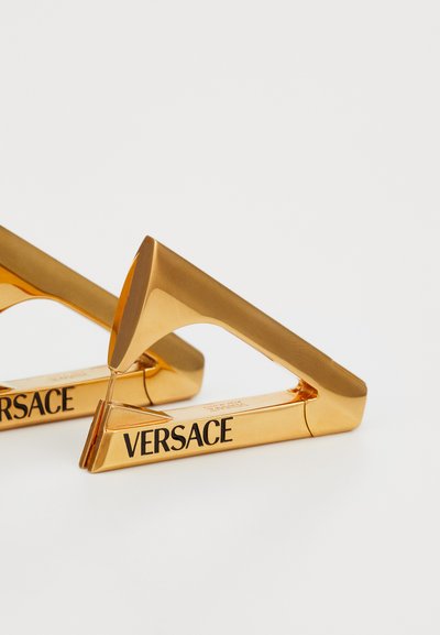 Auksinis metalinis spaustukas su trikampiu dizainu, turintis poliruotą paviršių ir juodą „VERSACE“ logotipą šone.