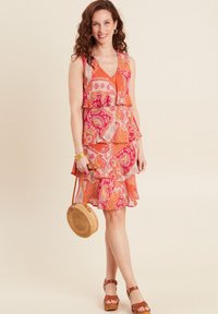 Ärmelloses Kleid mit Blumenmuster in Pink und Orange, mit geschichteten Rüschen; kombiniert mit einer runden, geflochtenen Tasche und braunen, schnürbaren Sandalen.