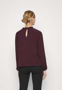Blusa lunga color marsala con dettaglio a apertura a chiave sul retro e scollatura arricciata. Tessuto morbido con una consistenza liscia e una vestibilità rilassata.