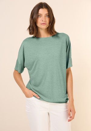 Top mint green a maniche corte realizzato in un tessuto morbido, con una vestibilità rilassata e piccoli dettagli decorativi. Indossato con pantaloni bianchi, mani in tasca.