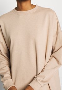 Sweatshirt beige surdimensionné avec des manches longues, col côtelé et tissu doux et texturé. Détail de couture subtil le long des coutures.