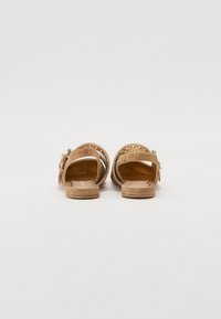 Sandali in suede beige con suola piatta, caratterizzati da un design a borchiette circolari sulla fascia superiore e chiusura con fibbia laterale.