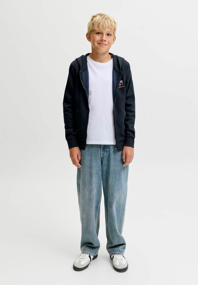 Garçon blond debout, portant un sweat à capuche noir zippé, un t-shirt blanc, un jean bleu ample et des baskets blanches, sur un fond clair uni.