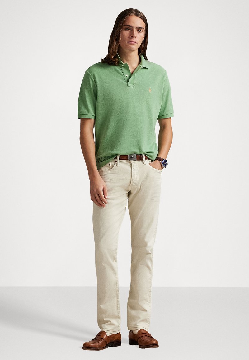 Groene polo in katoen met een klassieke kraag, gecombineerd met lichtbeige broek en leren schoenen in bruin; staande pose tegen een neutrale achtergrond.