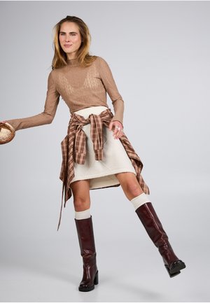 Žena s dlouhými vlasy, oblečená v hnědém pleteném topu, krémové sukni, kostkované košili uvázané v pase a tmavě hnědých kozačkách ke kolenům, držící malou pletenou tašku.