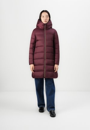 Bordeaux gewatteerde jas met een hoge kraag, ritssluiting en contrasterende rits, gecombineerd met brede blauwe jeans en glanzende zwarte schoenen.