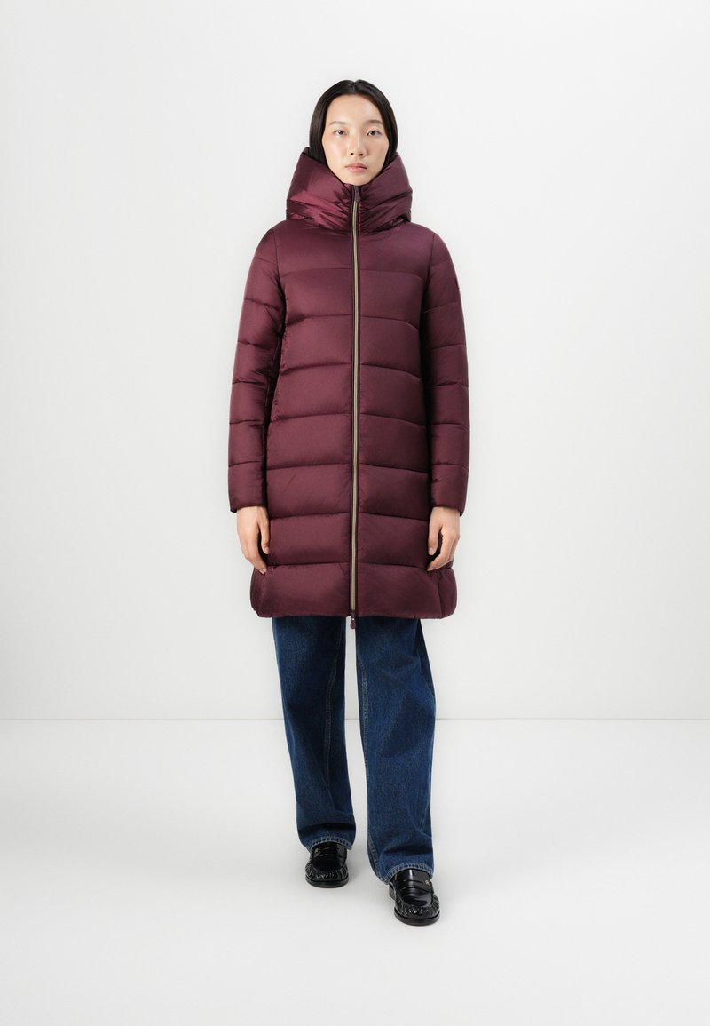 Burgundy vadderad jacka med hög krage, dragkedjestängning och kontrasterande dragkedja, matchad med vida blå jeans och blanka svarta skor.