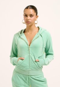 Felpa con zip verde menta realizzata in tessuto morbido, dotata di una tasca frontale, cappuccio regolabile e dettagli in zip dorata, abbinata a pantaloni coordinati.