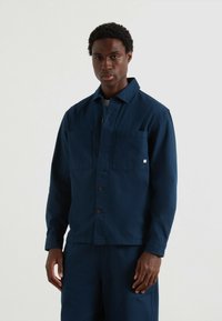 Uomo che indossa una giacca blu scuro con bottoni e due tasche sul petto, abbinata a pantaloni, in piedi di fronte a uno sfondo chiaro e uniforme.
