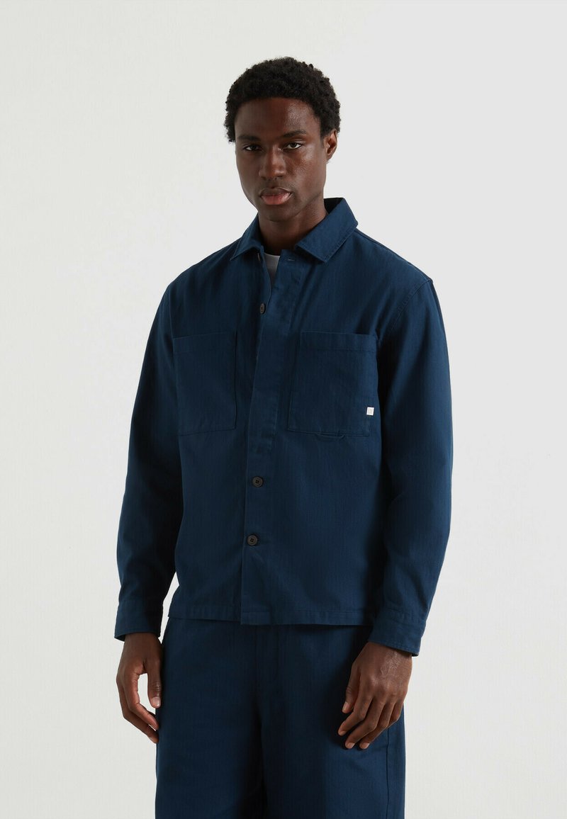 Uomo che indossa una giacca blu scuro con bottoni e due tasche sul petto, abbinata a pantaloni, in piedi di fronte a uno sfondo chiaro e uniforme.